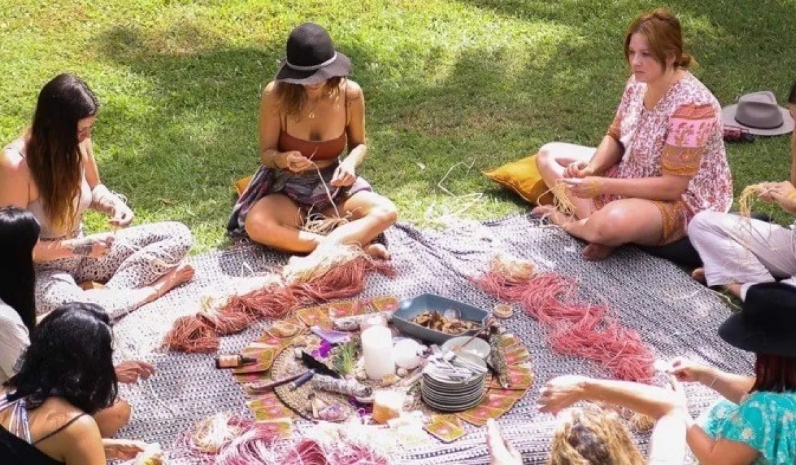 A group of women sit on grass and around a blanket upon which is thread and other items necessary for weaving.