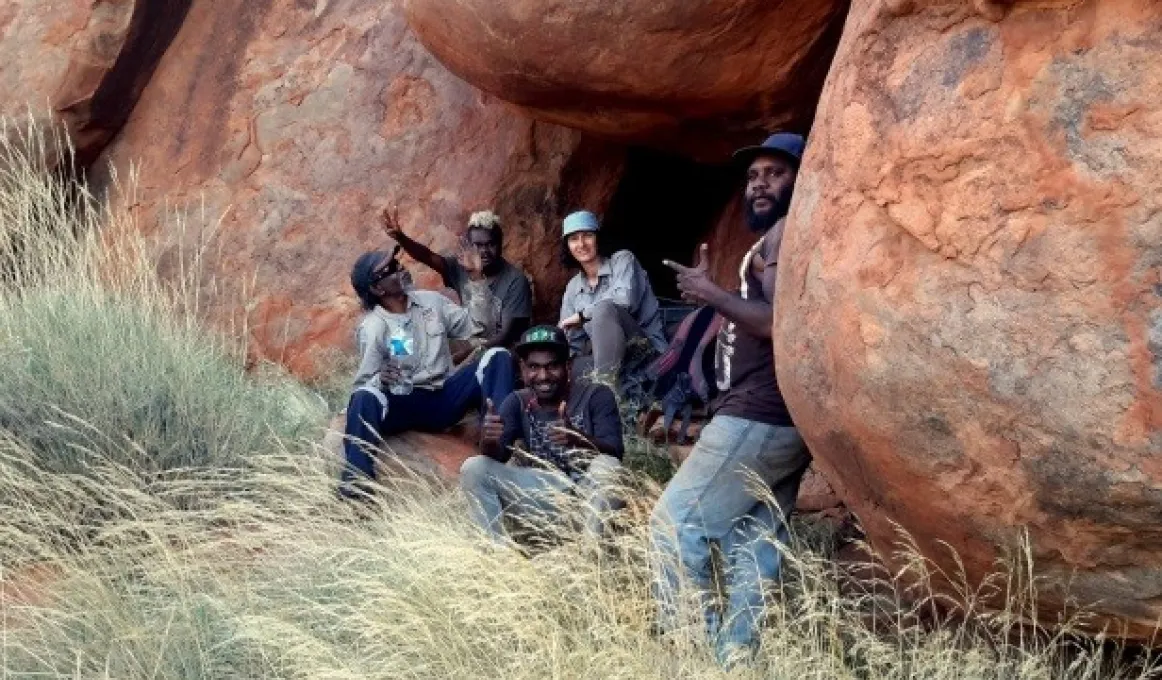 Group of people sitting under large rock formation. In the foreground is long grass.