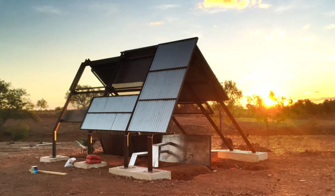 A structure of iron and metal sheeting stands on blocks of concrete. In the foreground is red soil and in the background are trees and a setting sun.