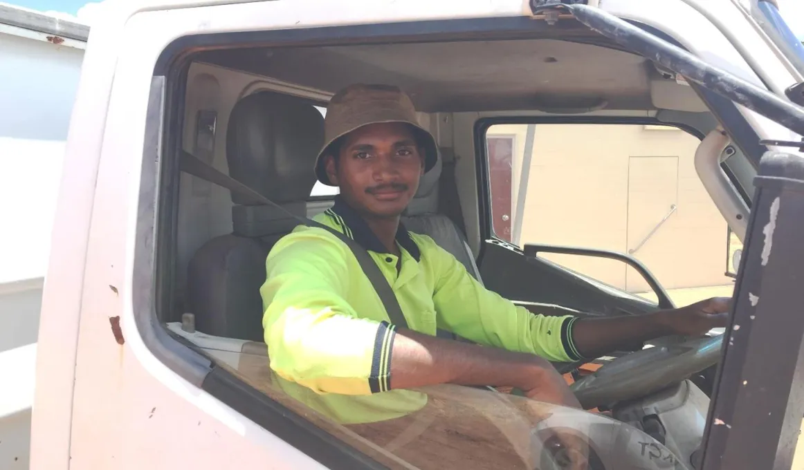 An Indigenous young man wearing work wear sits in the cab of a truck.