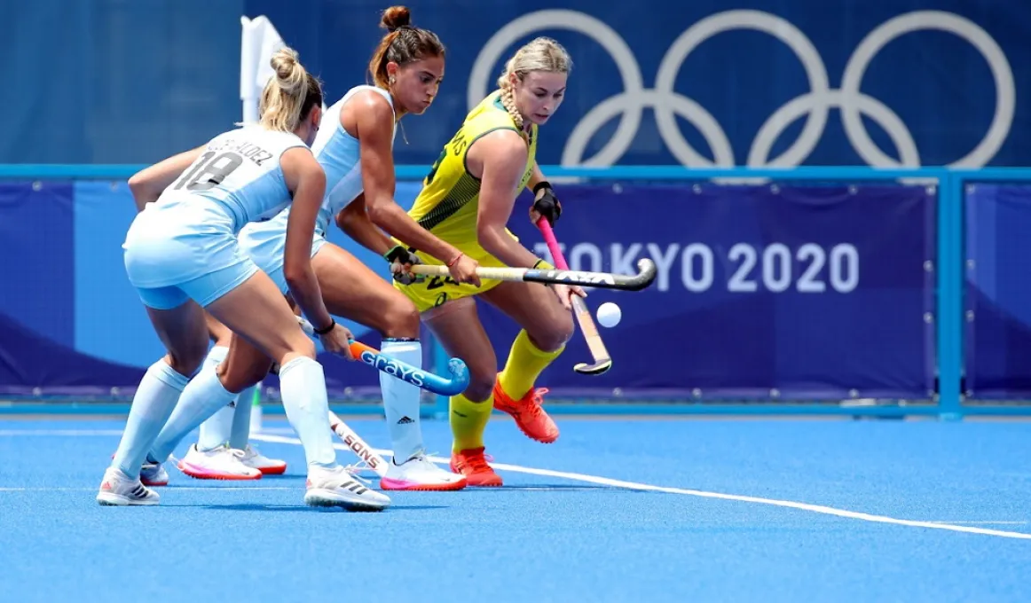 Three women, two dressed in blue uniforms and the other in yellow, hold hockey sticks and chase a small white ball on a blue surface. In the background are the words Tokyo 2020 and 5 rings overlaid in a line.