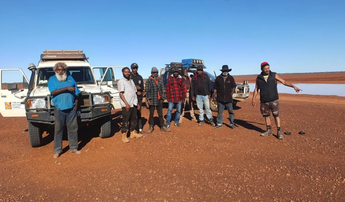 A group of men dressed in casual outdoor wear stand shoulder-to-shoulder on stony and ochre coloured soil. Behind them are two vehicles and in the background is a claypan and blue sky.