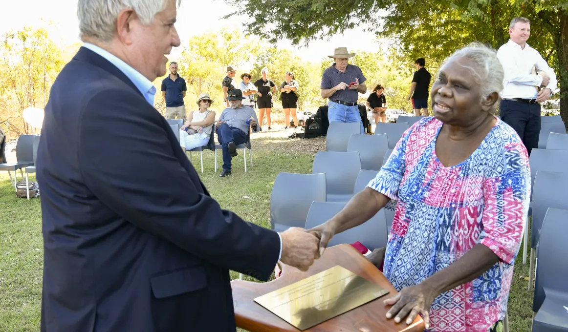 A man with grey hair and wearing a blue coat shakes hands with a woman with white hair and wearing a blue, pink and white dress. Under their hands is a podium and a gold engraved plate. In the background are chairs and people and trees.