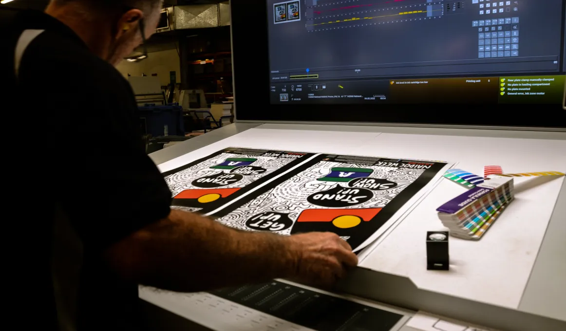 A man stands at a drafting table on which are two mainly black and white posters. To the right of the posters is a colour matching gauge. In the background is a large video screen.