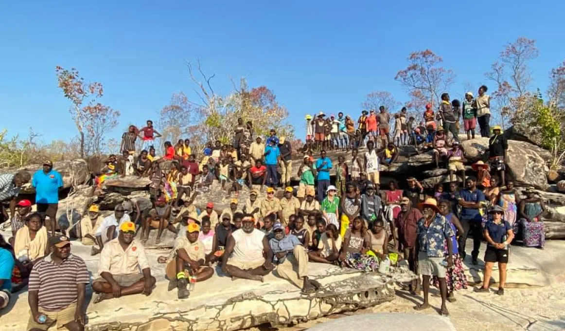 A large group of men, women and children wearing casual or work wear sit or stand on a rocky outcrop facing the camera. In the background are a few trees and a blue sky.