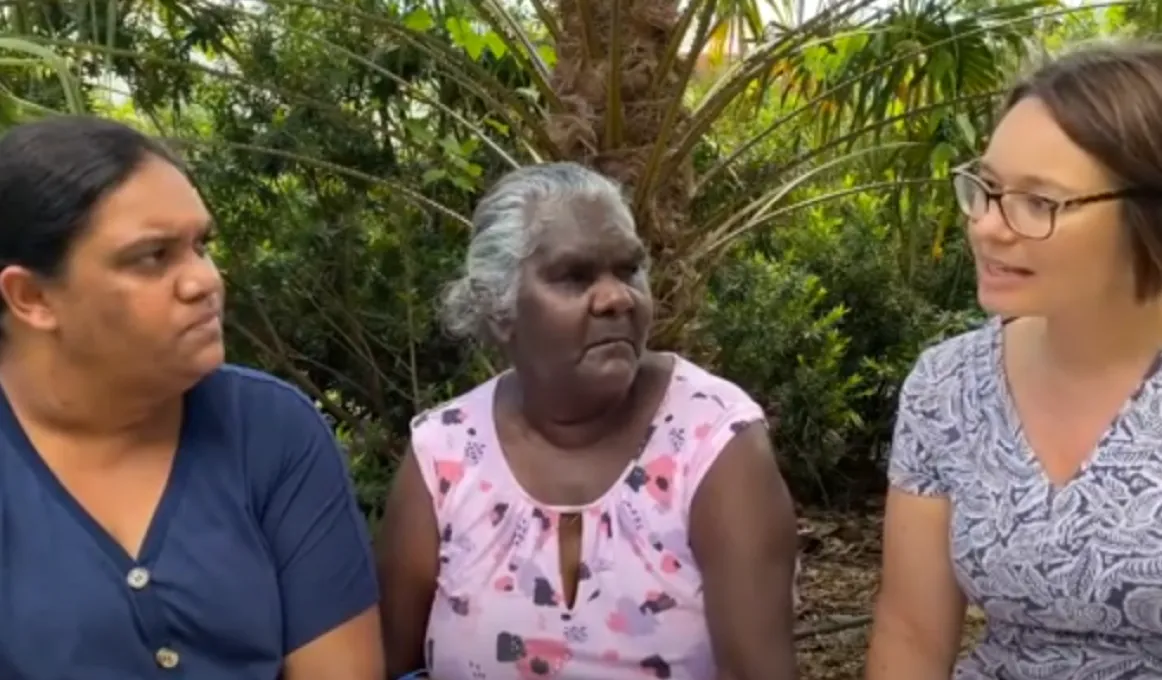 Three women sit next to one another in front of several trees and bushes in the background.