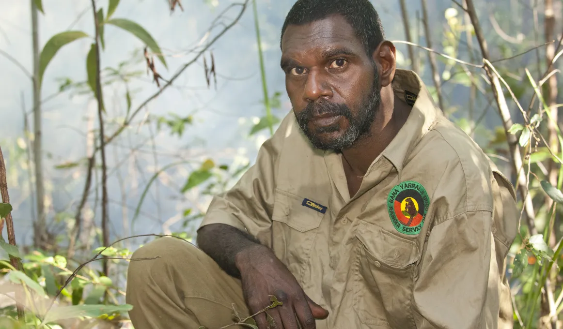 Indigenous man dressed in a ranger uniform sits in foreground with foliage in the background.