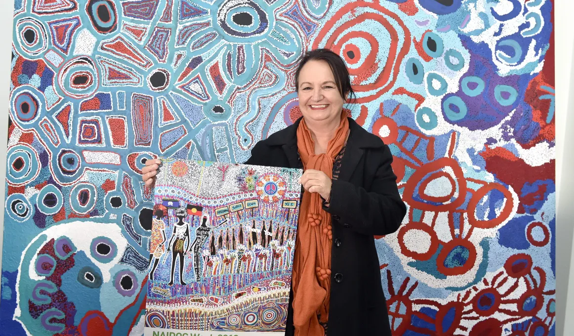 Cheryl Moggs, wearing a black jacket and orange scarf, holding the 2018 NAIDOC week poster ‘tarumunggie – woman.’ She is standing in front of a large, colourful canvas artwork.