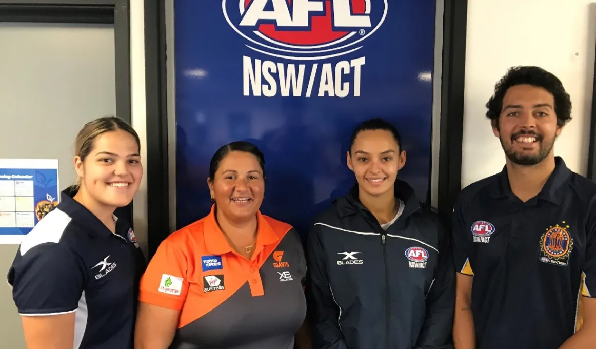 Three women and one man wearing AFL polo shirts stand in front of a large sign which says AFL NSW/ACT.