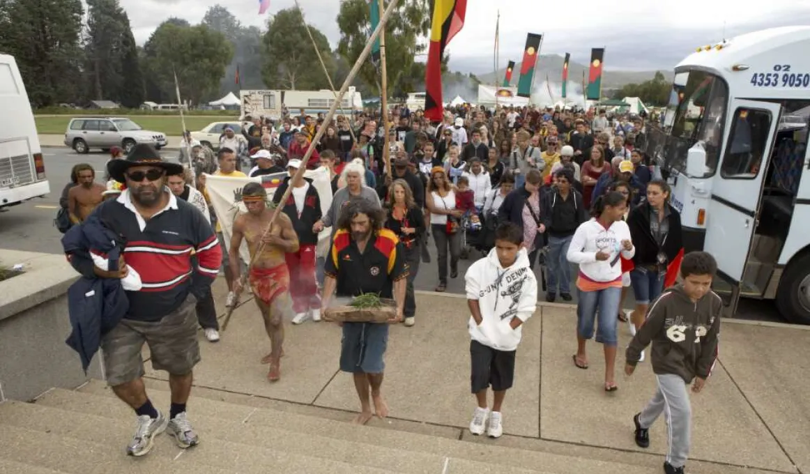 Large crowd of Indigenous men and women walking up stairs away from the Aboriginal Tent Embassy bearing Aboriginal flags coloured black on top and red on the bottom with a yellow circle. One man in a black shirt with red and yellow panels carrying a piece