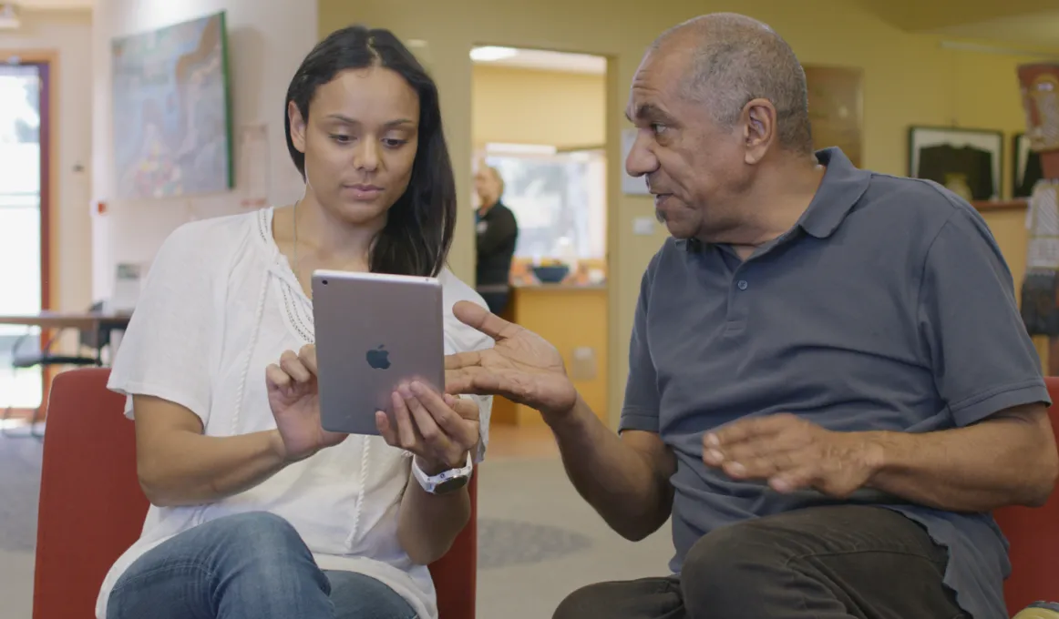 A young adult woman sits on a red chair holding a small tablet device. She is showing the screen to an older man who is gesturing to the device with his hand. They look calm and relaxed.