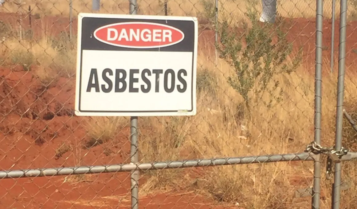 A sign reading “Danger Asbestos” attached to a gate with a background of red dirt and dry grass.