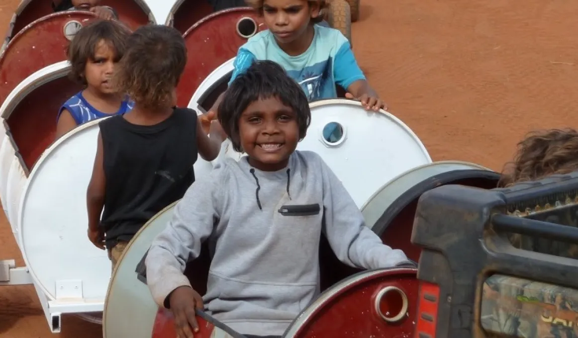 Young Aboriginal children sit in and stand next to a series of barrels mounted on wheels.