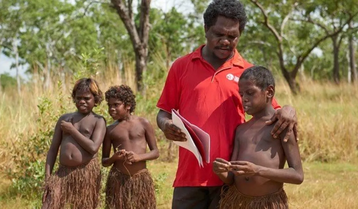 Two Aboriginal boys in grass skirts stand at left and behind an Aboriginal man in a red shirt and boy in a grass skirt in the foreground. The man holds some papers on which he and the third boy are focussed. In the background is long grass and trees.