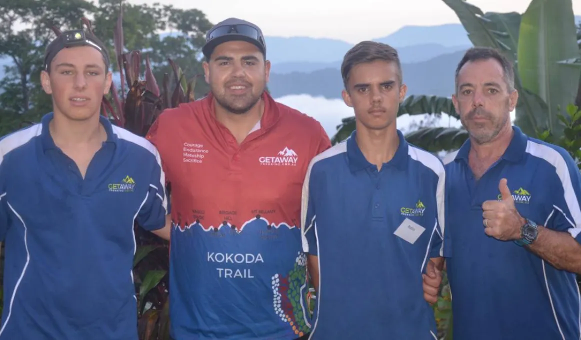 Two adult males and two young men dressed in mainly blue shirts, stand arm in arm in front of tropical foliage atop a mountain. In the background is mountain terrain and low-lying cloud.