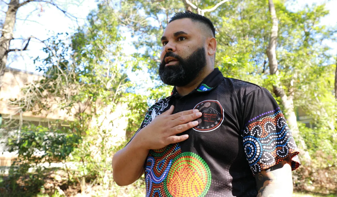 A man stands in the shade on a sunny day, with trees behind and around him.  He has his hand on his heart and is wearing a shirt with an Aboriginal artwork print on it.