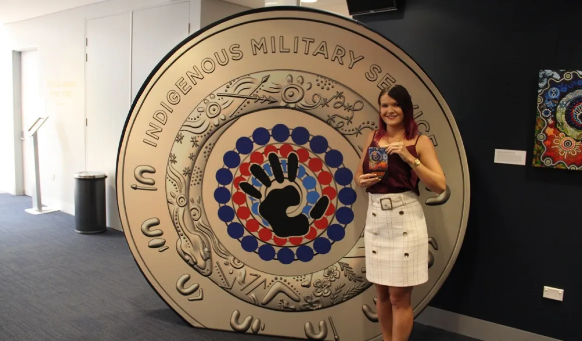 A woman in pale skirt and dark top stands next to a large replica of a coin, which features a hand in the middle of concentric circles, made up of black, red and blue dots. The coin is gold in colour. In the background are walls and a carpeted floor.