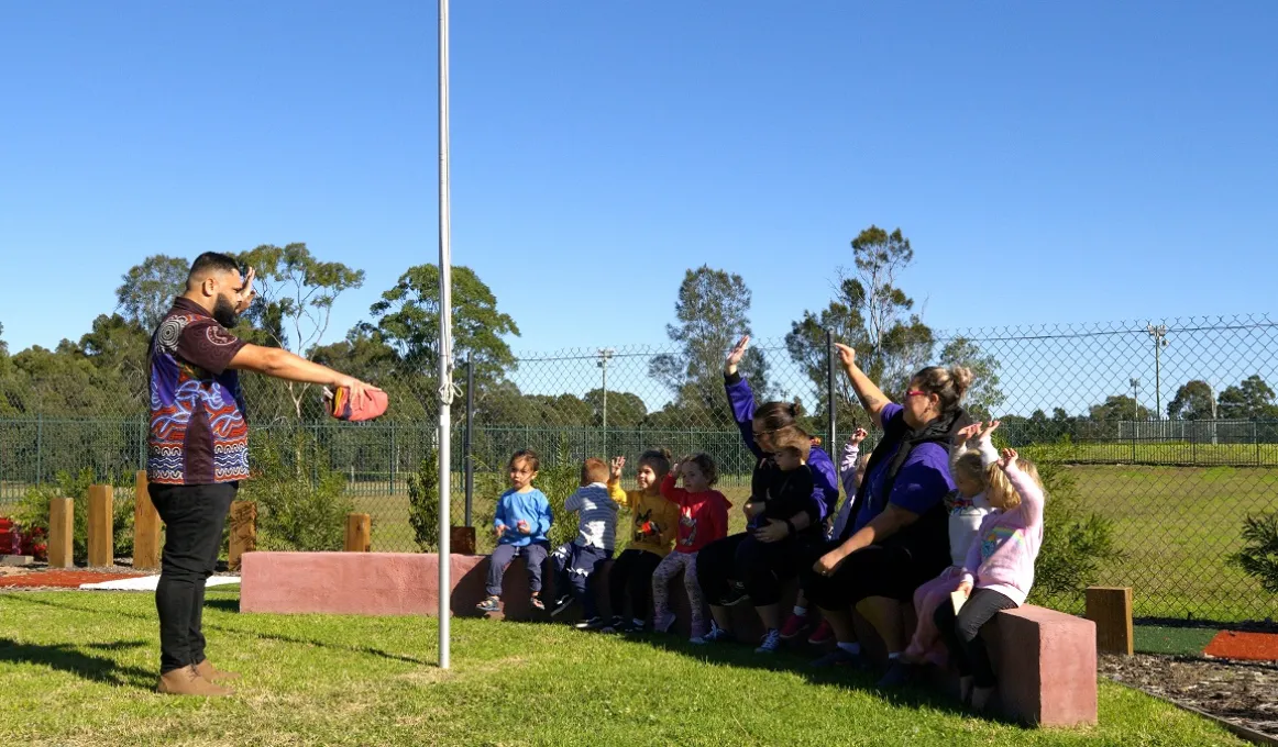 Group of children and adults sit on a low wall. In front is a grassed area on which is a pole. On the other side of the pole stands an Aboriginal man. In the background is a fence and trees and more grass.