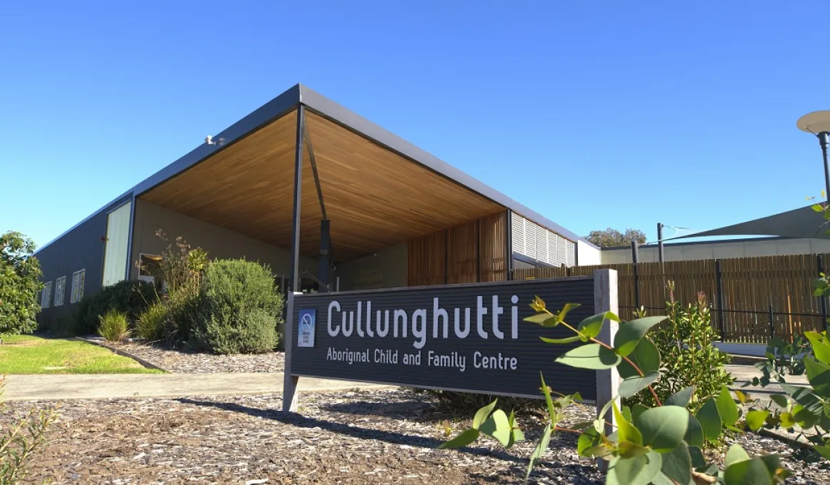 Large sign in foreground with the words Cullunghutti Aboriginal Children and Family Centre. In the background is a building with shrubs and around it. Behind the building is a sail and a clear blue sky.