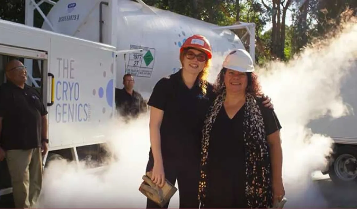 2 women are standing outside on a sunny day. They are wearing hardhats and smiling at the camera. In the background are 2 men, standing next to a large vehicle. White smoke is rising from the ground.