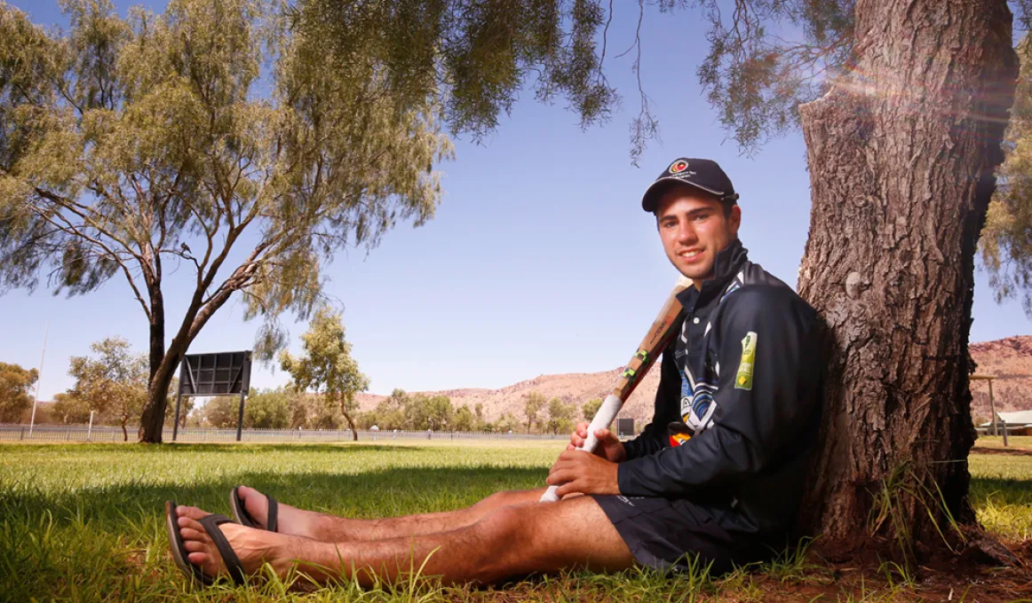 Young Aboriginal man in dark blue hat, shirt and shorts sits under a tree leaning up against the trunk while holding a cricket bat. In the background is a grassed area, a sign, many trees and a large brown hill.