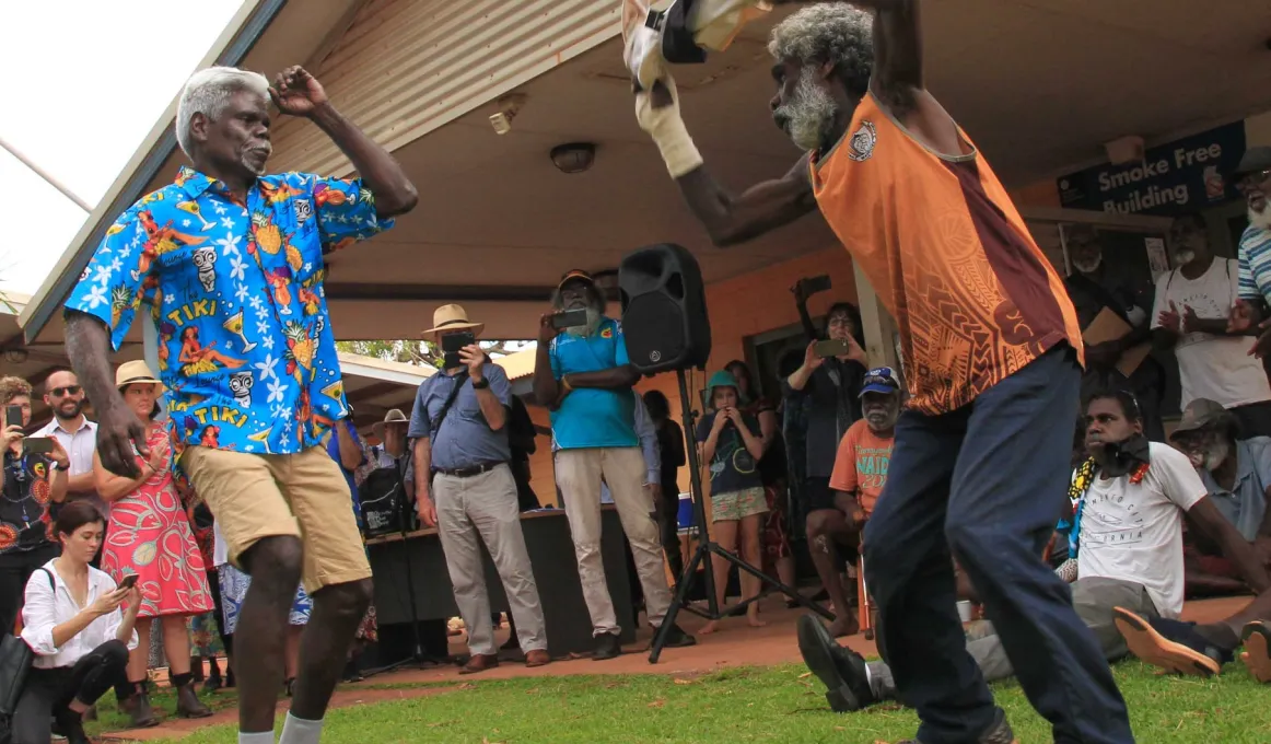 Two men dancing in front of a crowd. The dancers are wearing colourful tops and dancing with material in their hands. They are dancing in front of a crowd of Indigenous and non-Indigenous peoples