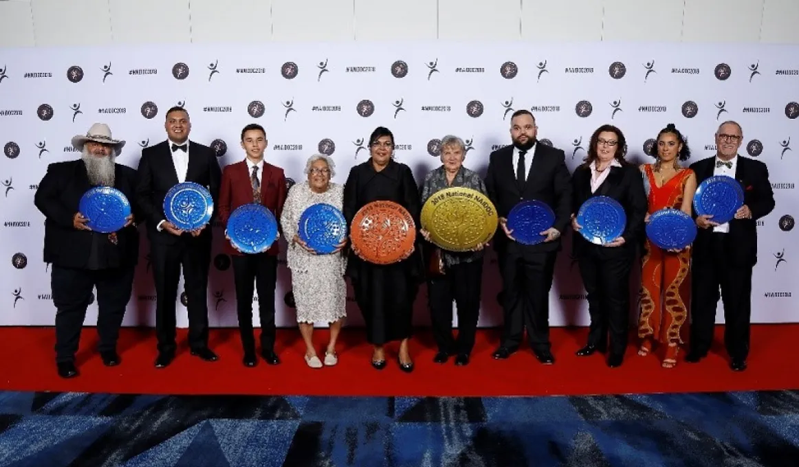 12 award winners stand in a row with a logo photo wall behind, on the NAIDOC 2018 red carpet