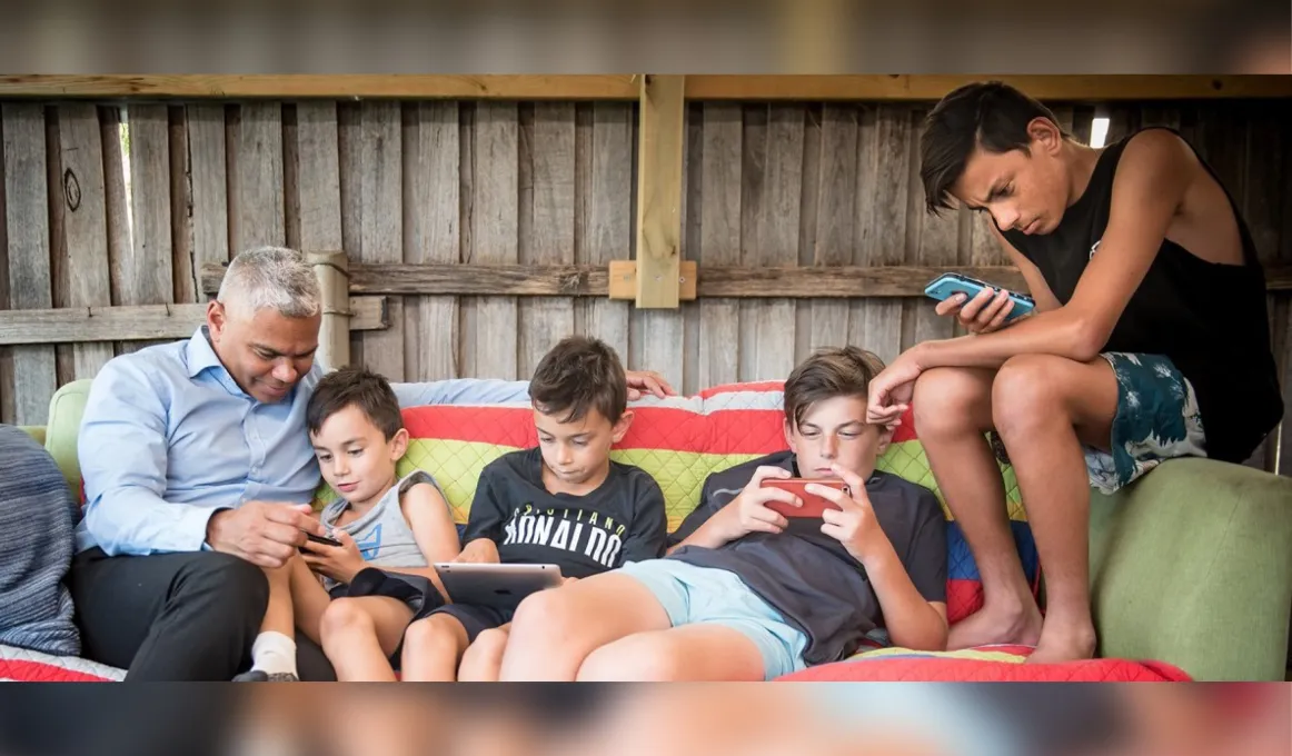 Indigenous man in shirt and long pants sits on a couch with boys dressed in shorts and t-shirts or singlets. In the background is a wood paling fence.