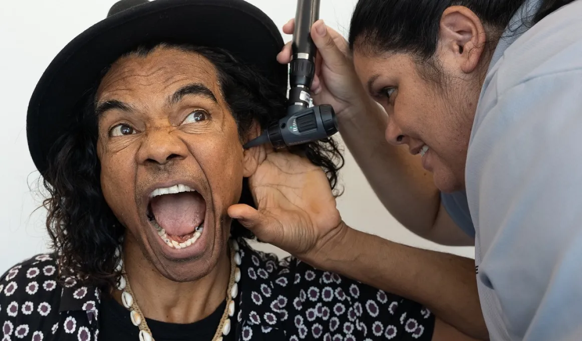 An Indigenous man in hat and black and white shirt opens his mouth and holds his ear as an Indigenous nurse applies an instrument to his ear.