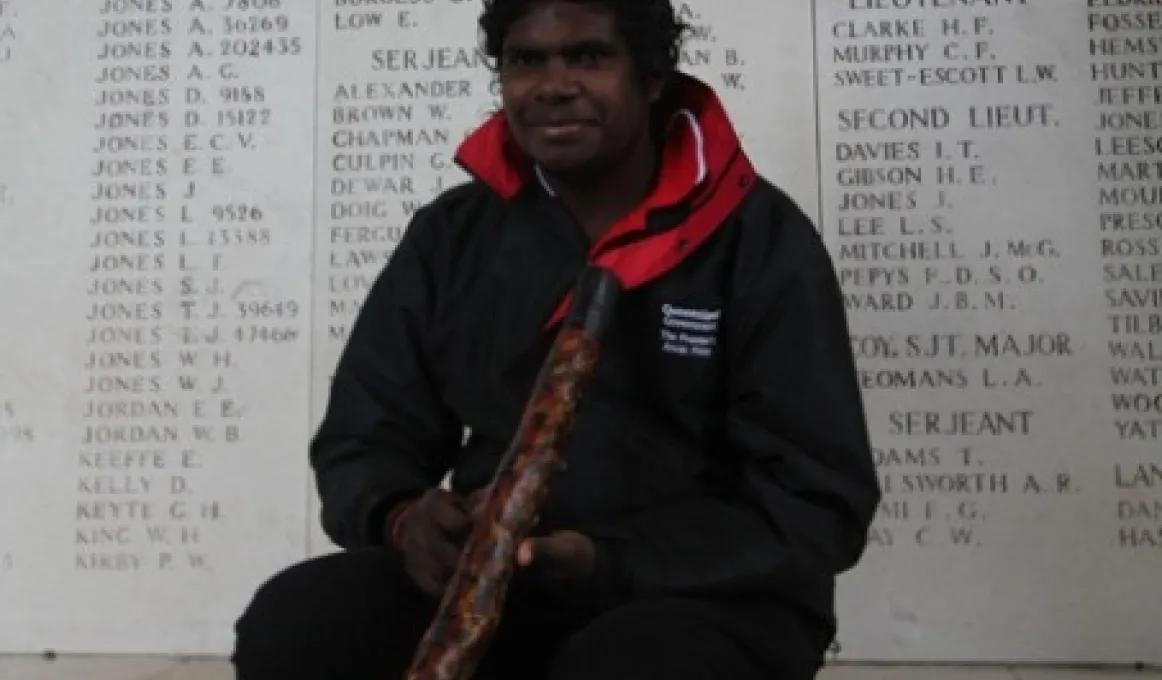 Elijah Douglas, holding his didgeridoo, sits in front of a wall of remembrance.