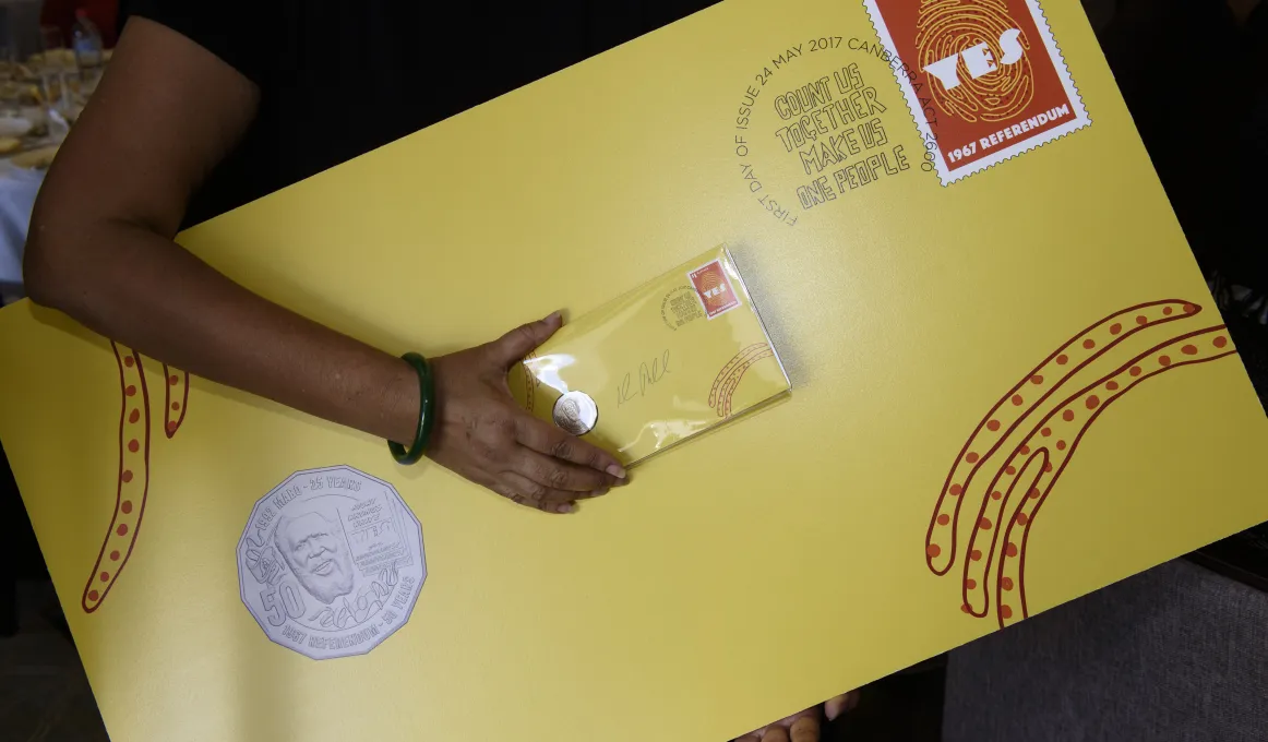 Indigenous person holding a large yellow envelope with ‘Yes’ stamp and new Eddie Mabo coin attached along with a regular size envelope of the same design. The envelope includes some Indigenous design.