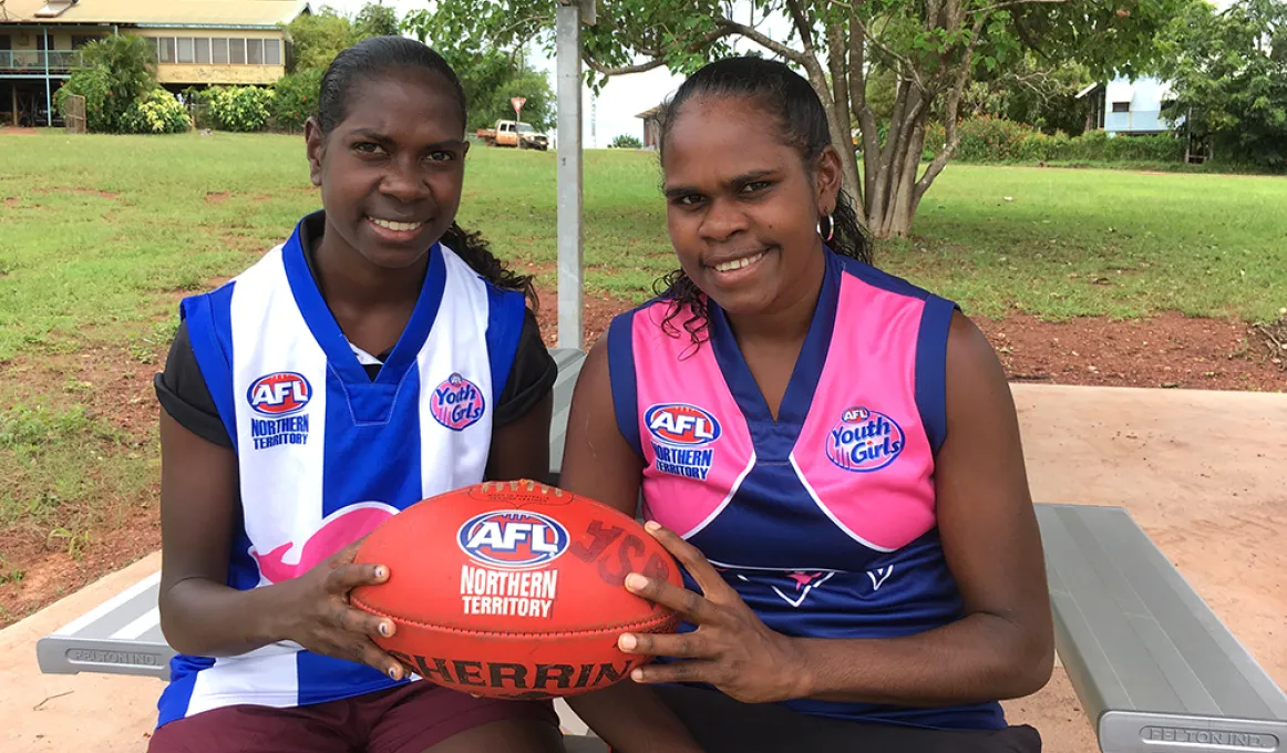 Two young Aboriginal women sit at a picnic table, holding an AFL football in their hands. One wears a blue and pink football jumper, the other wears a blue and white striped football jumper. In the background is a grassed area, trees and buildings.