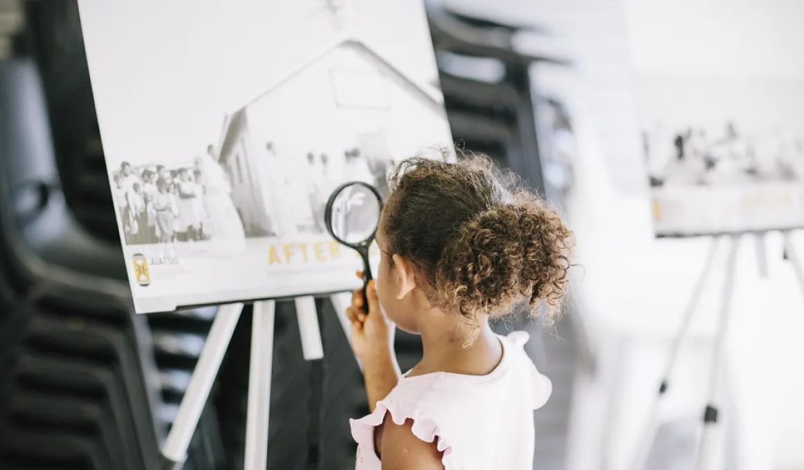 A young girl with her brown hair drawn back holds a magnifying glass up to a photo of people next to a building.