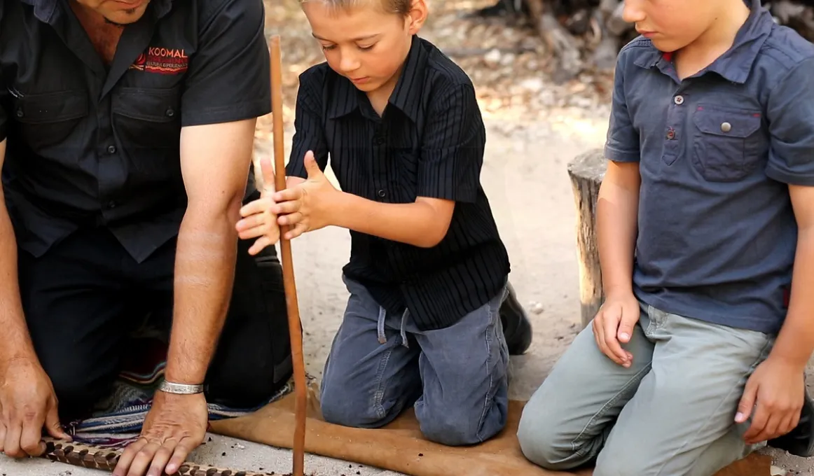 A man and two boys kneel on a skin on the ground. The man holds a long stick on the ground. One boy holds another stick at right angles from above and spins the stick in his hands. The other boy looks on.