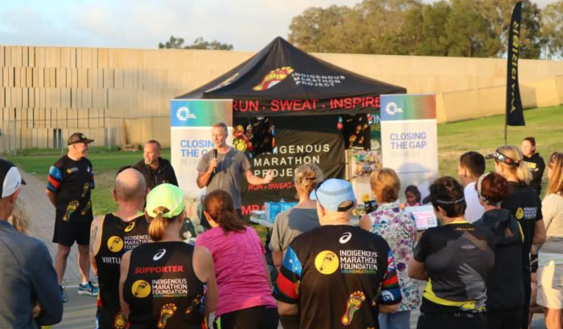 Group of people with backs to us dressed in running wear look at man in grey shirt holding microphone standing in front of a small tent with banners either side saying Closing the Gap and on the tent are the words Run Sweat Inspired.