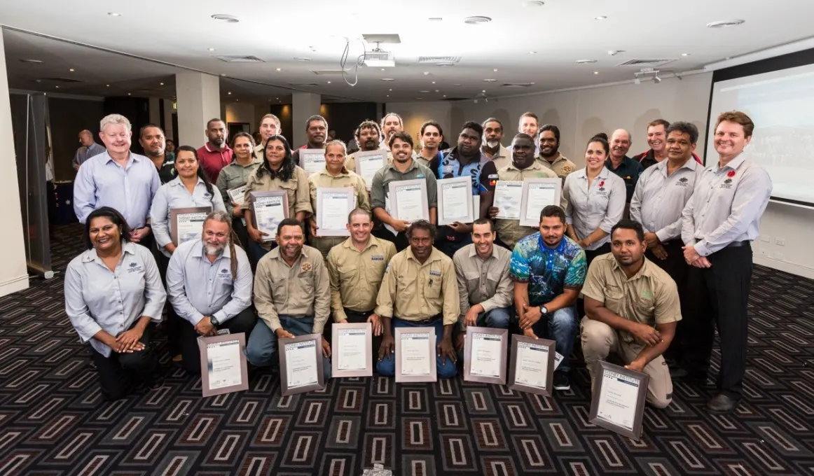 Large group of Indigenous and non-Indigenous people in a carpeted room facing the camera, some dressed as rangers are holding certificates of achievement.