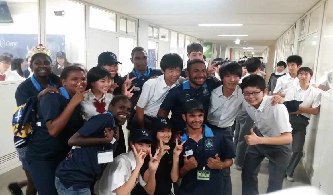 A group of Japanese students in white shirts arm in arm with Aboriginal and Torres Strait Islander students in blue shirts. In the background are more students and classrooms.