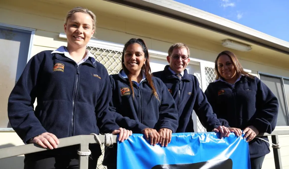 Four young adults wearing blue jumpers standing in front of a building and hanging onto a rail.