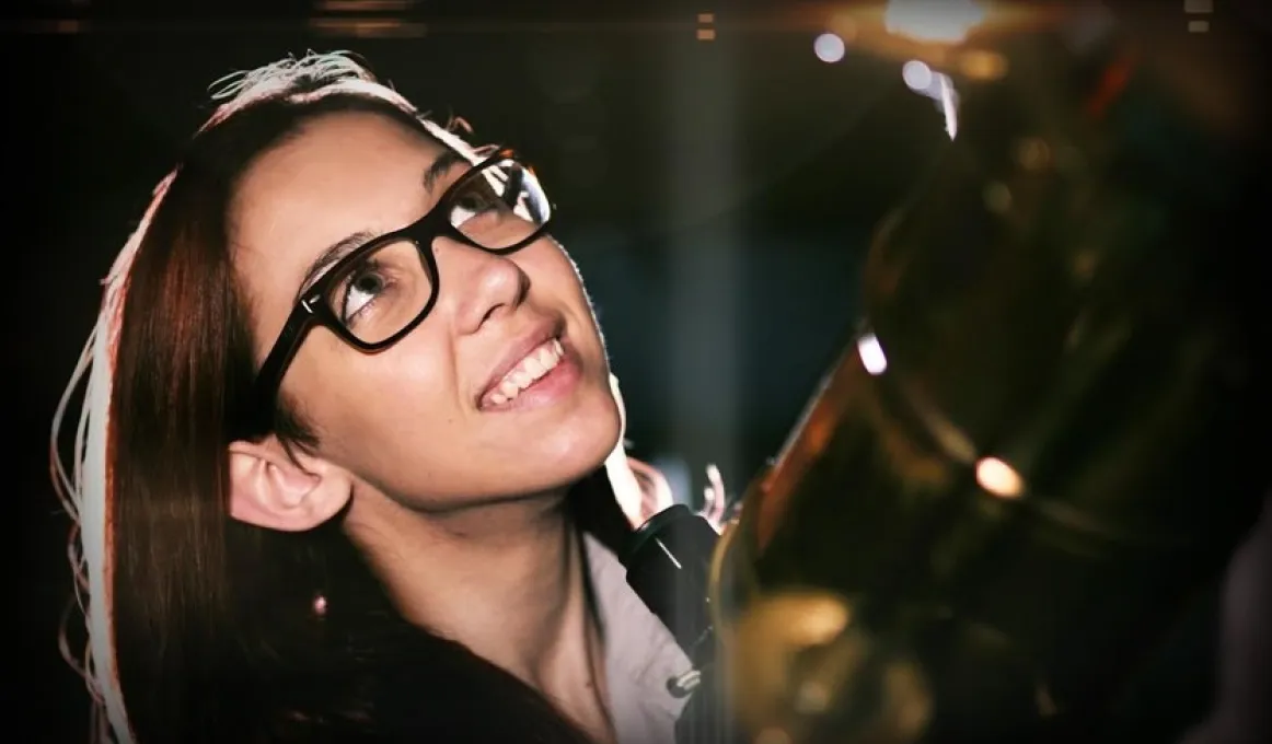 Photo of a young woman, Kirsten Banks, looking up at the sky next to a telescope.