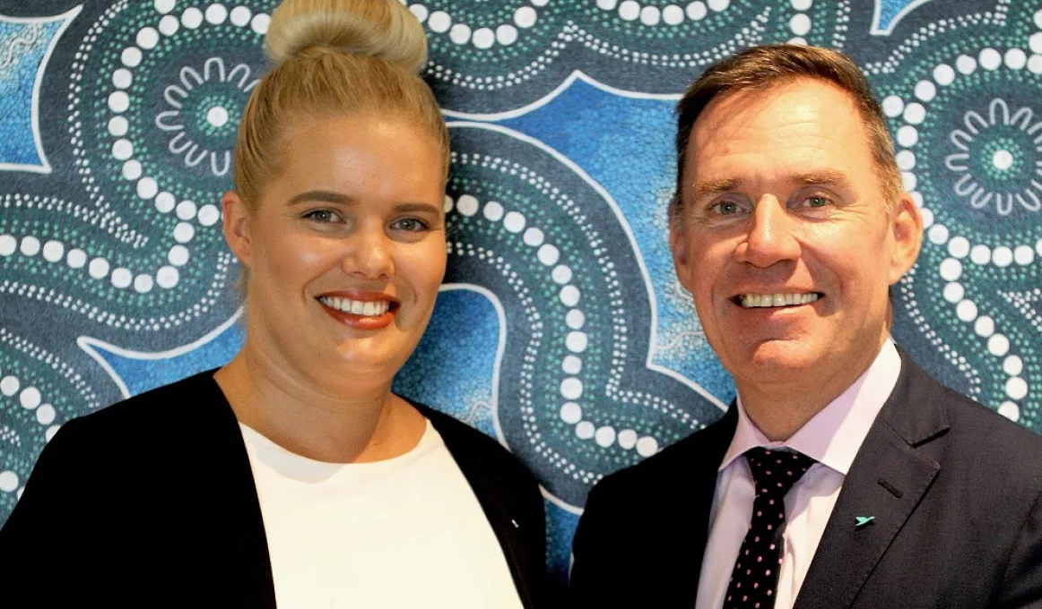 Indigenous woman in white top and black jacket with non-Indigenous man dressed in suit and tie stand in front of wall covered with blue, white and green Indigenous designs