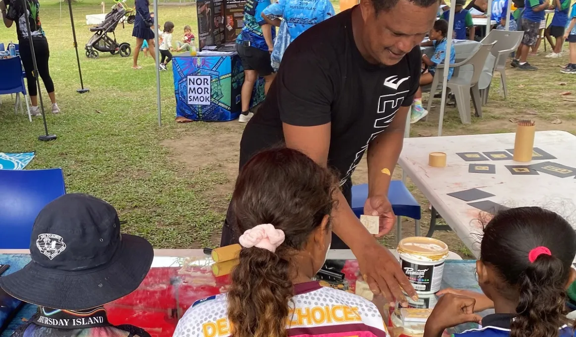 A man and three children work at a table which has paint pots on it. In the background are more tables, gazebos and people.