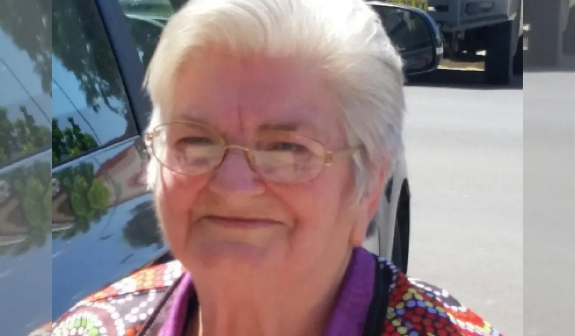 An elderly Aboriginal woman in a colourful polo shirt standing outside next to a car.