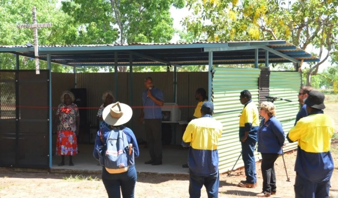 Group of Indigenous and non-Indigenous people standing in and outside of a green shed with a cross on top.