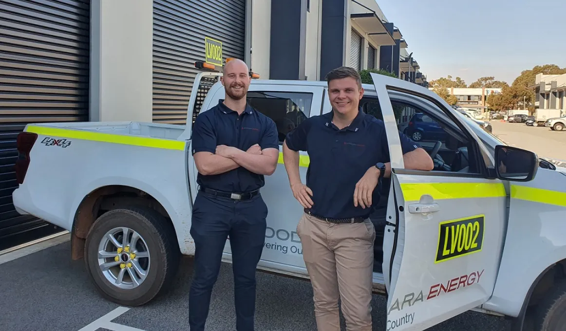 Two men stand side by side in front of a work vehicle. Both are dressed in work wear. Behind them is an industrial shed with multiple doors and more buildings and cars further in the background.