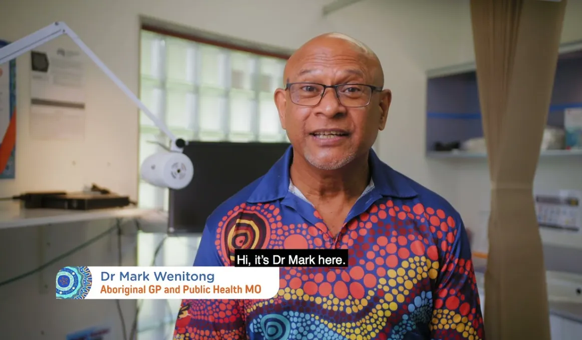 A man sitting in doctor’s surgery wearing a brightly coloured polo shirt and glasses. Words read: Doctor Mark Wenitong Aboriginal GP and Public Health MO. Hi it’s Dr Mark.