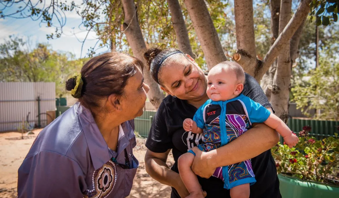 Two Aboriginal women smile at an Aboriginal child held by one of them. The child wears a blue jumpsuit with Aboriginal designs on the front. In the background is a flower bed, two fences and trees.