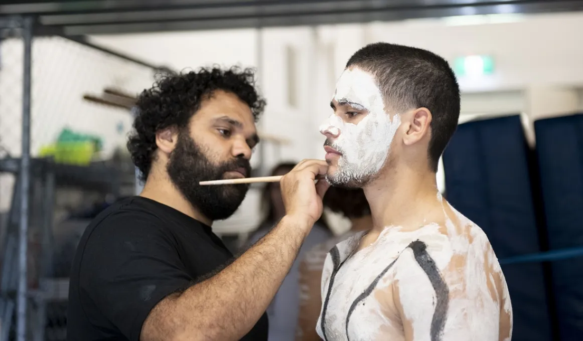 A man in black t-shirt is painting the upper body of another man. He has paint on his face, shoulders and chest.