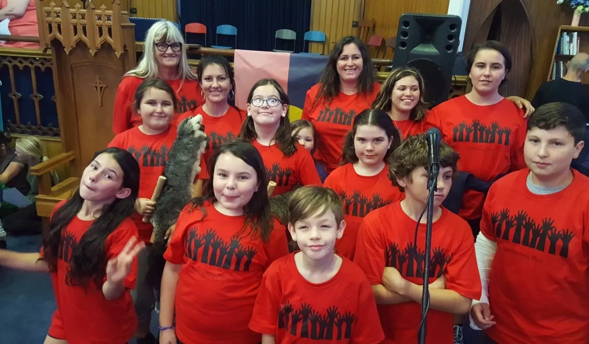 Two adult women and twelve children stand in a room behind a microphone, all wear red t-shirts with a graphic of raised hands.