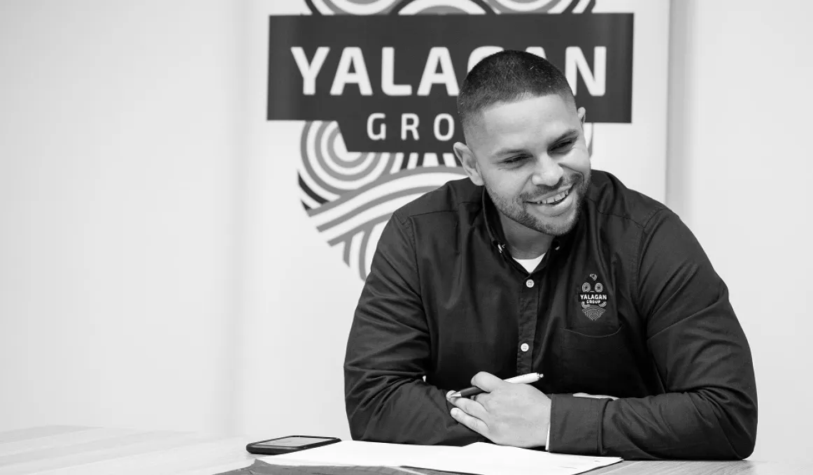 Young adult man in dark shirt sits at a table. In the background is a logo including the words Yalagan Group.