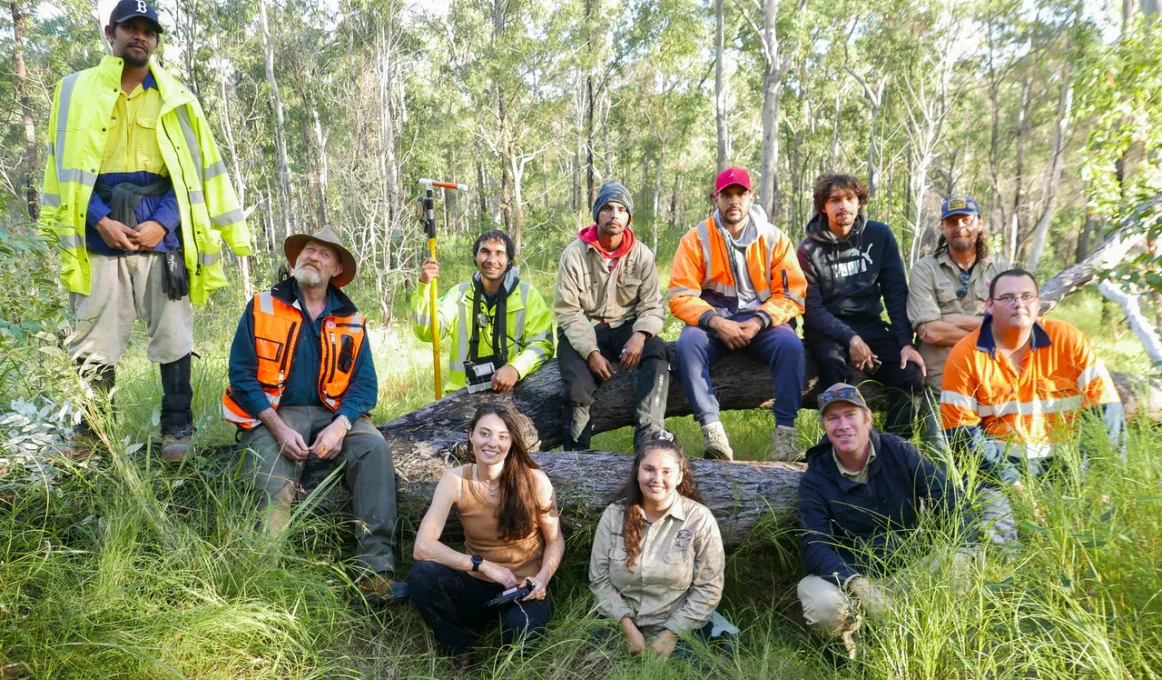 A group of people sit on or in front of a fallen tree and face the camera. Most are dressed in workwear. In the background are trees and grass. In the foreground is more grass.